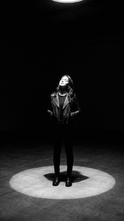 Nova Rai standing in a circle of light on a dark floor, looking up